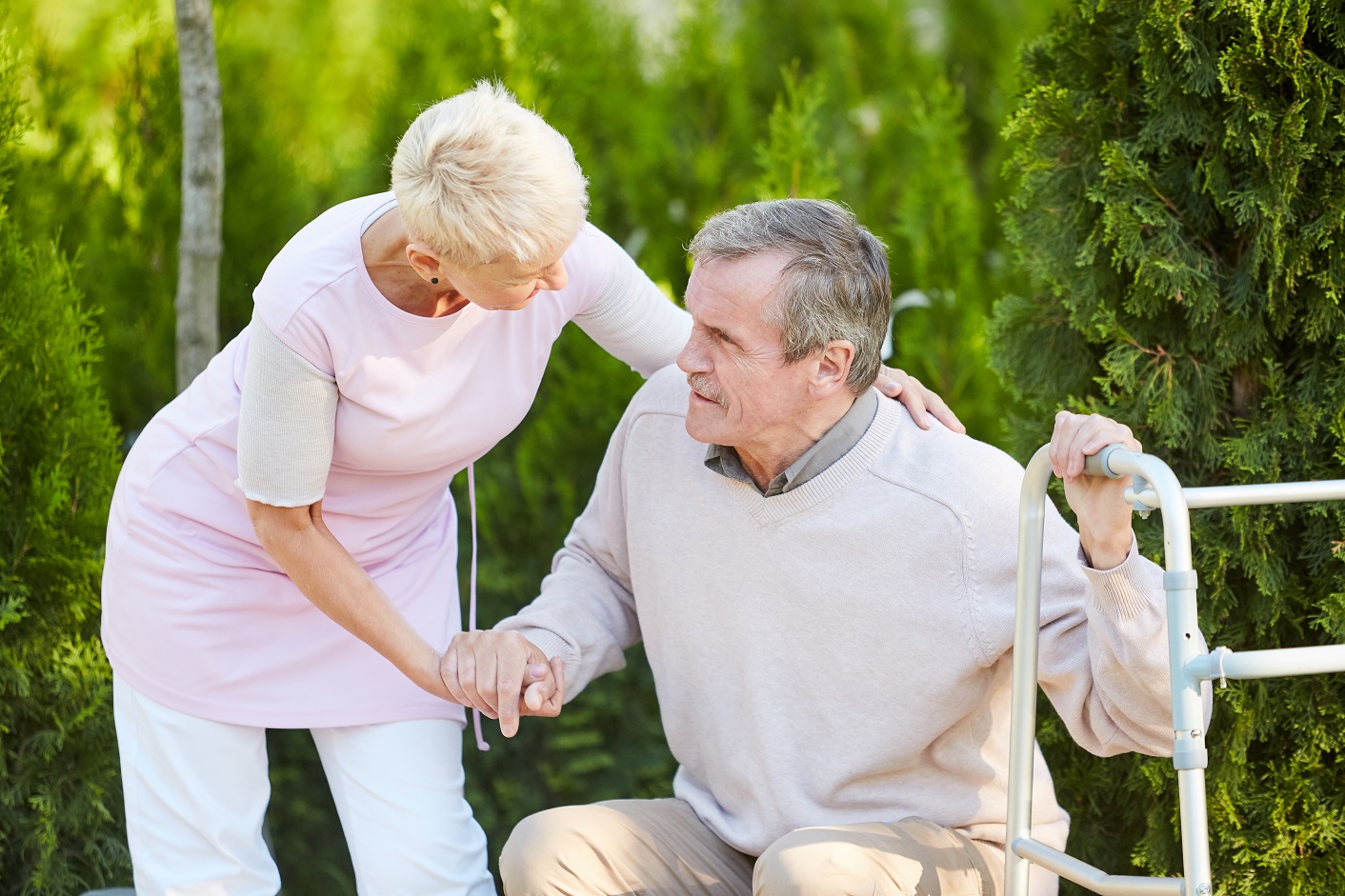 Portrait of caring nurse helping senior man get up from park bench in rehabilitation center, copy space
